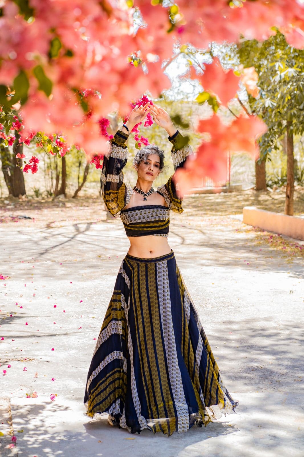 Indigo and ochre block-printed lehenga set with a patterned crop-top and striped pleated skirt — full-length silhouette beneath a bougainvillea canopy.