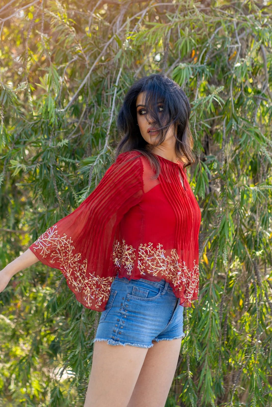 Sheer crimson chiffon pleated top with crimson-and-gold thread embroidery along a scalloped hem — bell sleeves, styled over denim shorts in willow foliage.