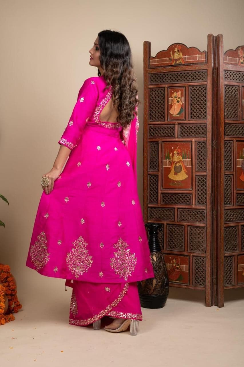 Fuchsia silk anarkali with gold gota patti motifs and a sheer pink dupatta — flared hem with bold floral embroidery, photographed beside a hand-painted Rajasthani screen.