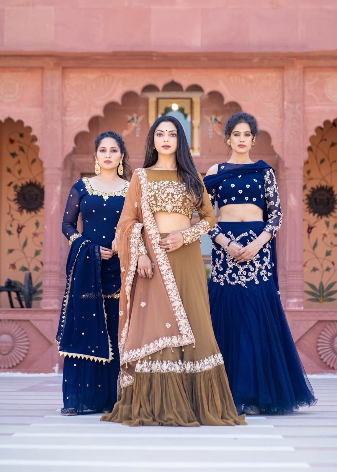 Trio portrait — sapphire blue mermaid lehenga, caramel and ivory zardozi set, and indigo mesh lehenga — photographed beneath the cusped arches of a Jaipur haveli forecourt.