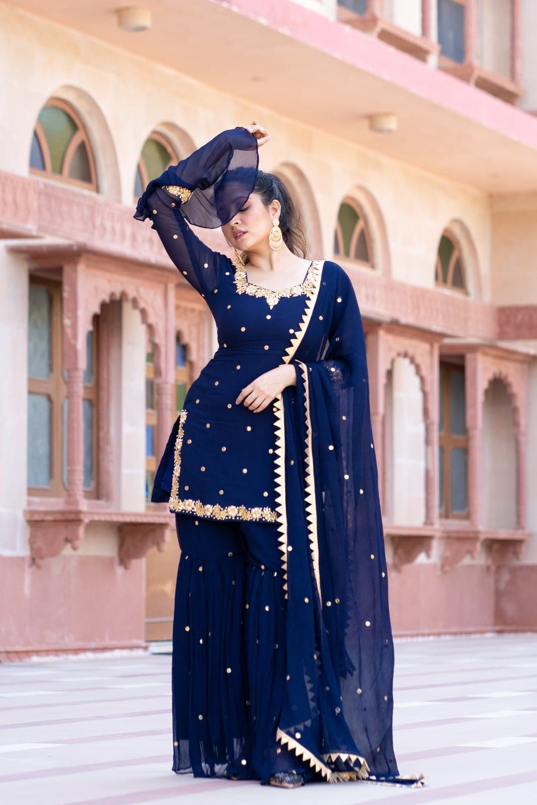 Navy chiffon kurta and gharara with gold mukaish dot embroidery and a zig-zag gold-bordered dupatta — photographed against the sandstone arches of a Jaipur fort terrace.