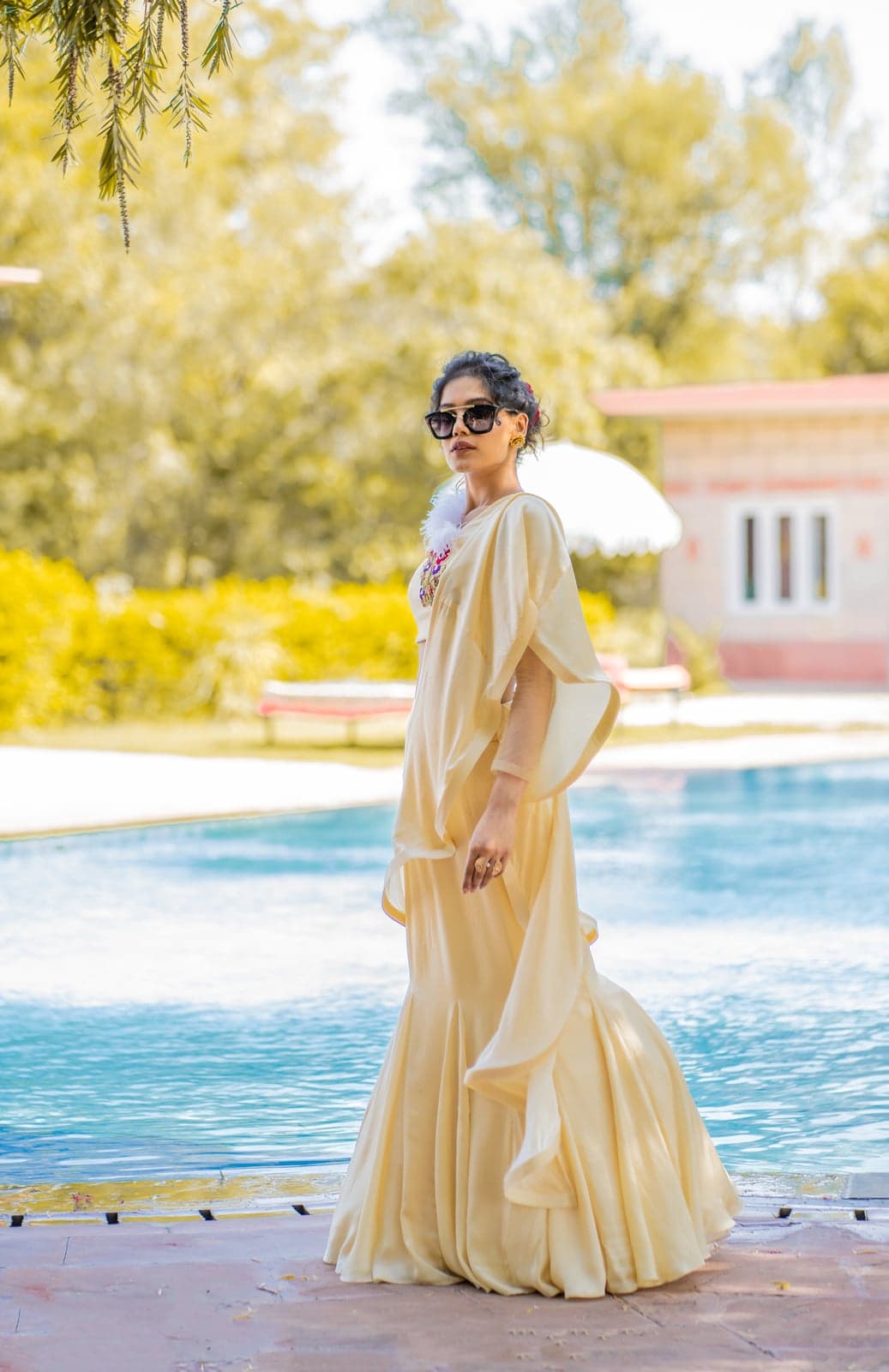 Champagne ivory drape gown with a sculpted mermaid hem and floral-yoke cape — late-morning light by the pool.