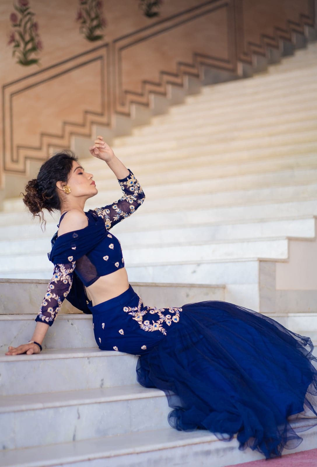 Sapphire blue lehenga seated study — off-shoulder bodice with zardozi sleeves, tulle mermaid skirt cascading across marble steps.