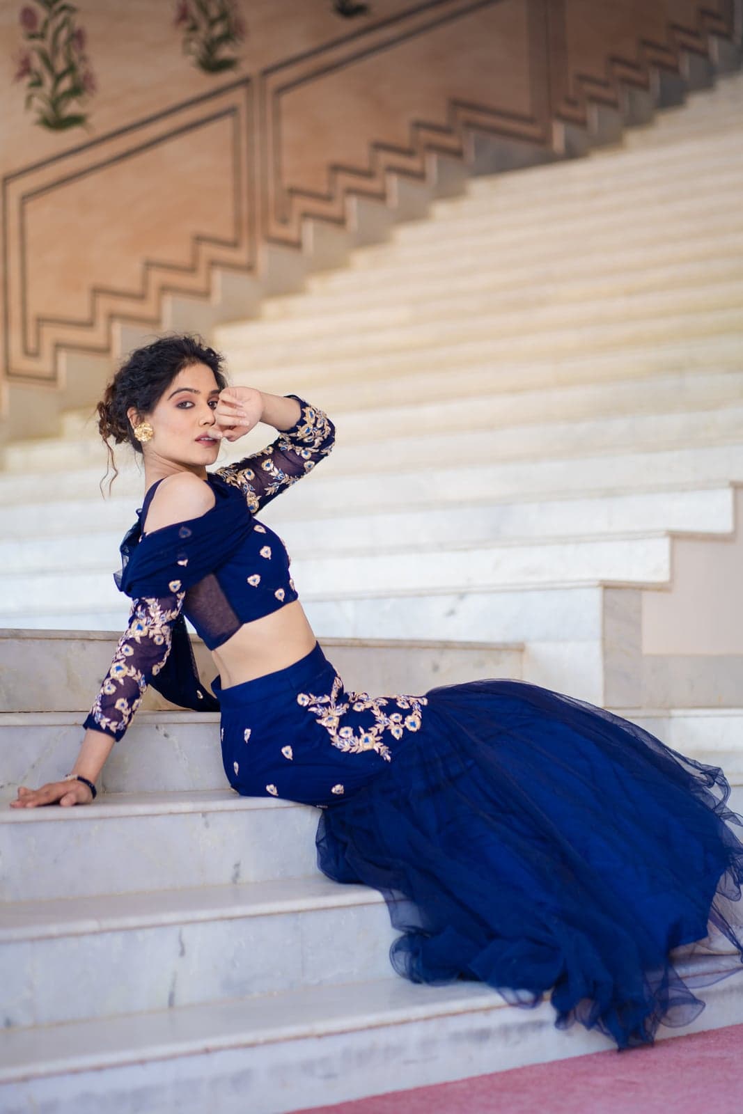Sapphire blue lehenga in profile silhouette — off-shoulder bodice, embroidered sleeves, tulle skirt photographed across a Jaipur palace staircase.
