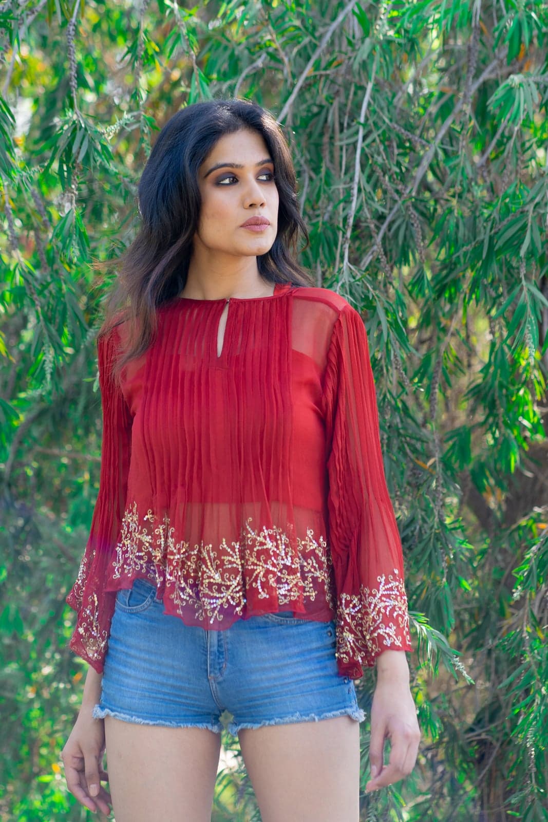 Sheer crimson chiffon top with crimson-and-gold floral hem embroidery — close portrait in willow foliage.