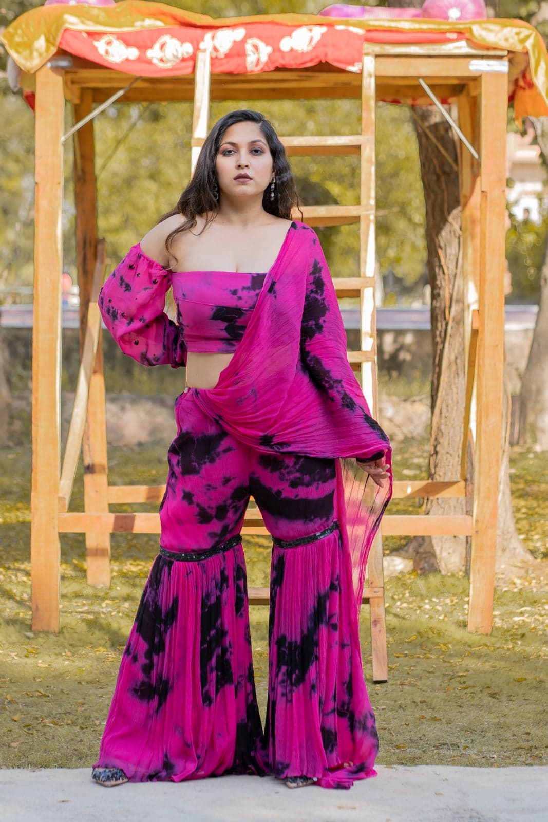 Magenta and black tie-dye gharara — single-figure portrait under the marigold mandap, soft daylight filtering through the fabric.