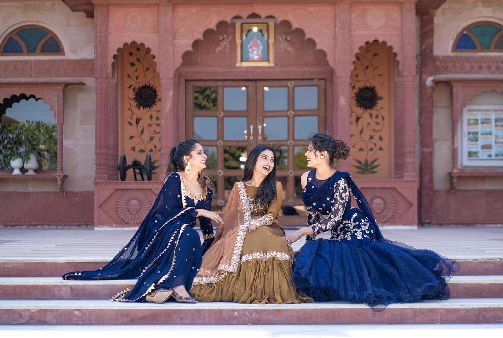 Group portrait on the haveli forecourt — sapphire blue mermaid, caramel zardozi, and indigo mesh lehengas posed together in conversation.