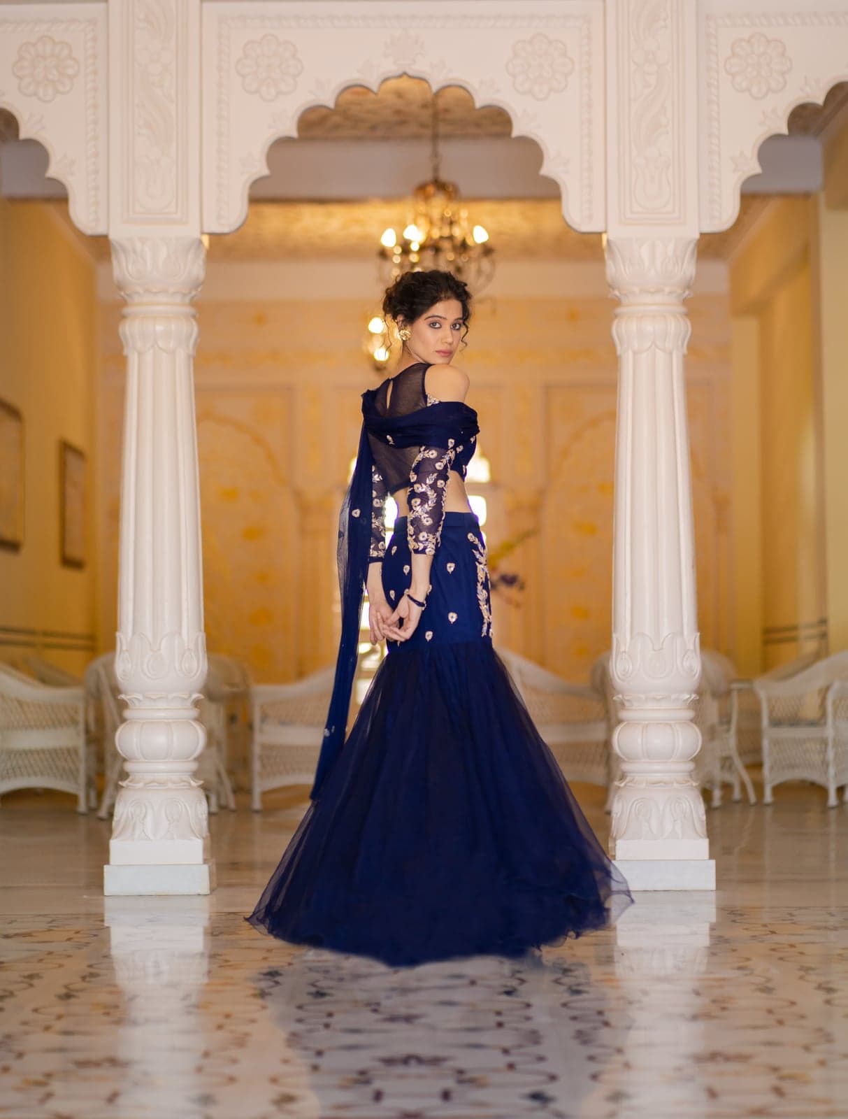 Sapphire blue mermaid lehenga from behind — full silhouette study, palace pillars and chandelier framing the train.
