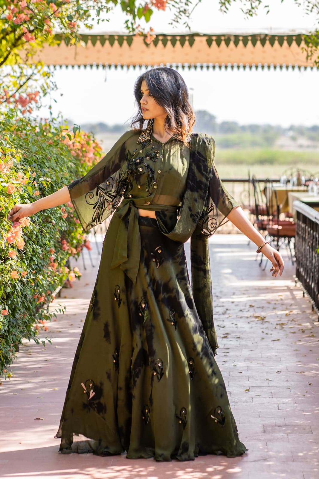 Olive green sequined sharara set — full-length view with tie-dye drape skirt, photographed amid a leafy courtyard pergola.