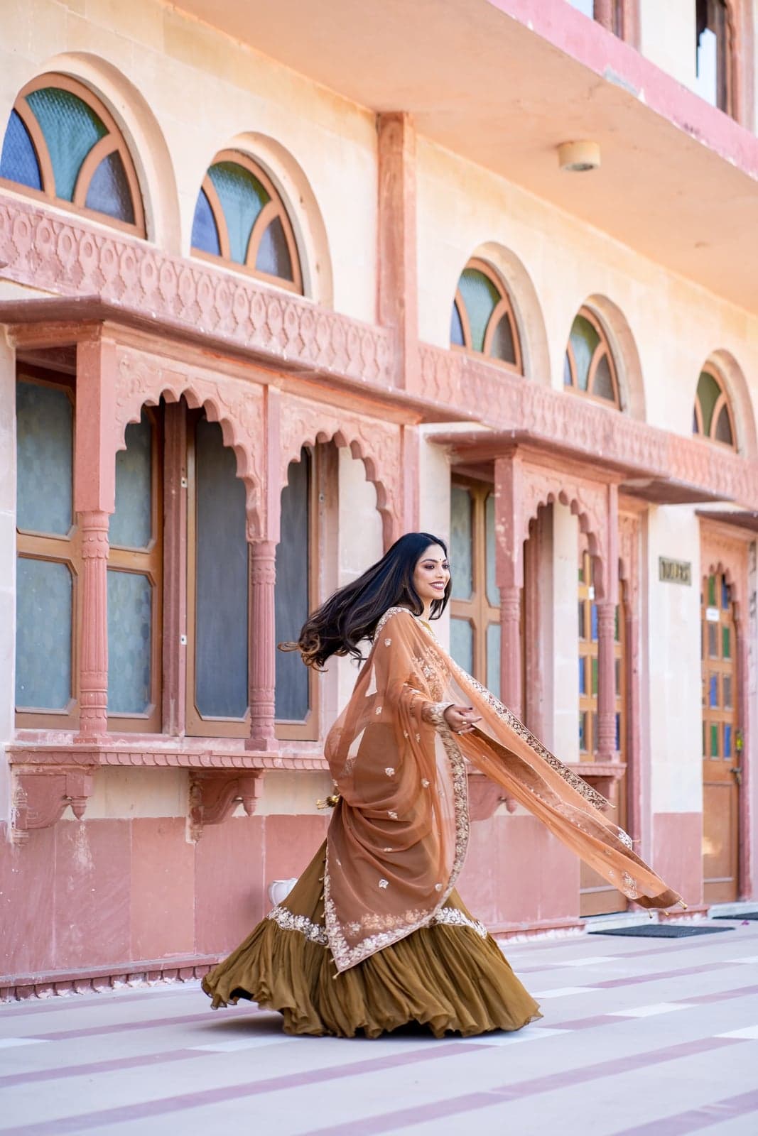 Caramel brown silk lehenga in motion — gold-embroidered organza dupatta caught mid-twirl against the arched sandstone facade of a Rajasthani haveli.