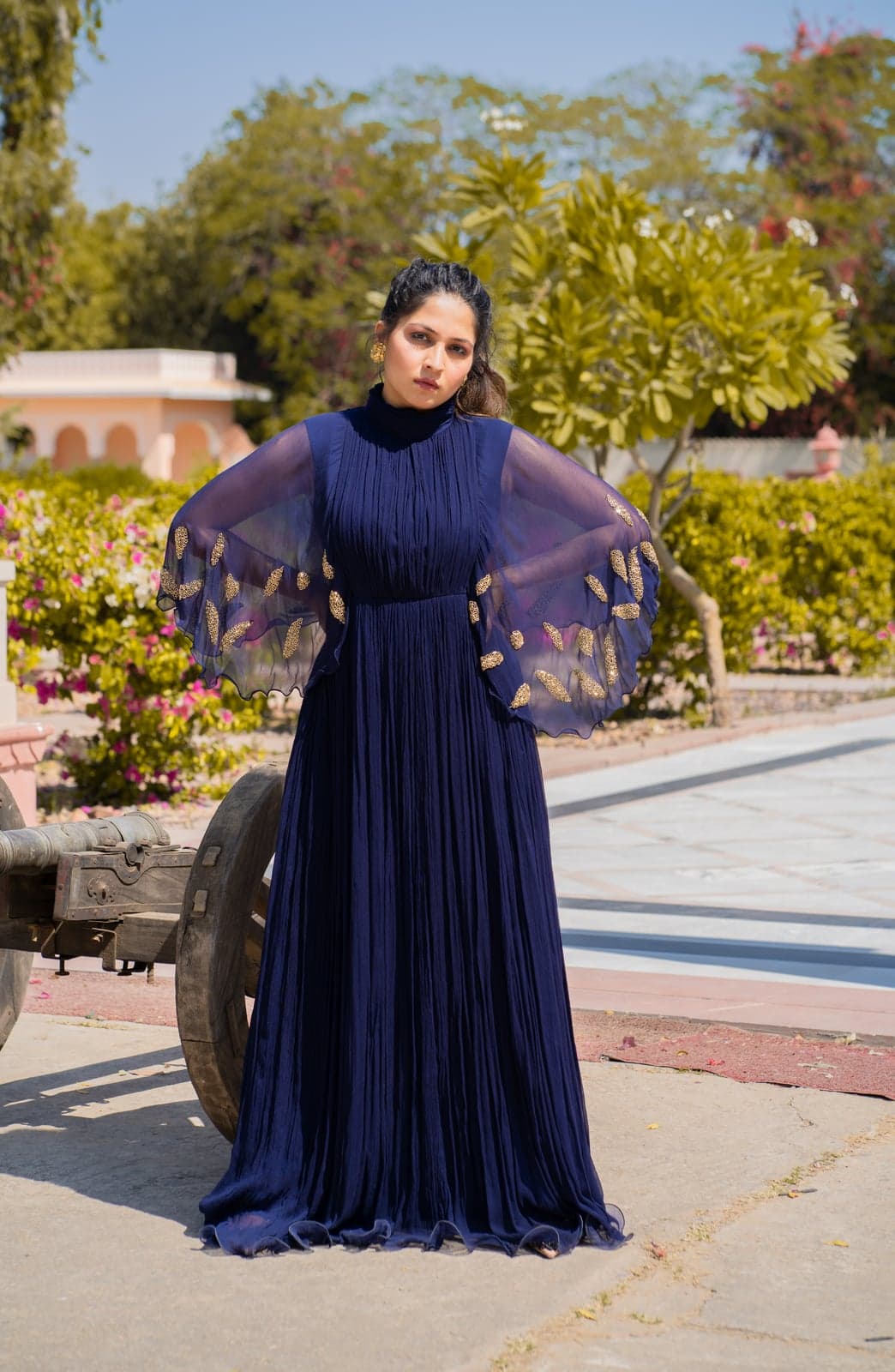 Navy pleated chiffon gown — butterfly-cape sleeves with gold leaf embroidery, full-length portrait in a Jaipur garden beside a wooden cart wheel.