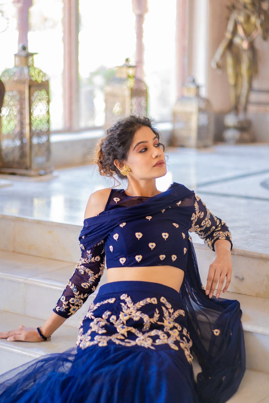 Sapphire blue lehenga reclining detail — gold-on-blue zardozi embroidery, sheer veil over the bodice, mehrab arches of a Jaipur palace beyond.