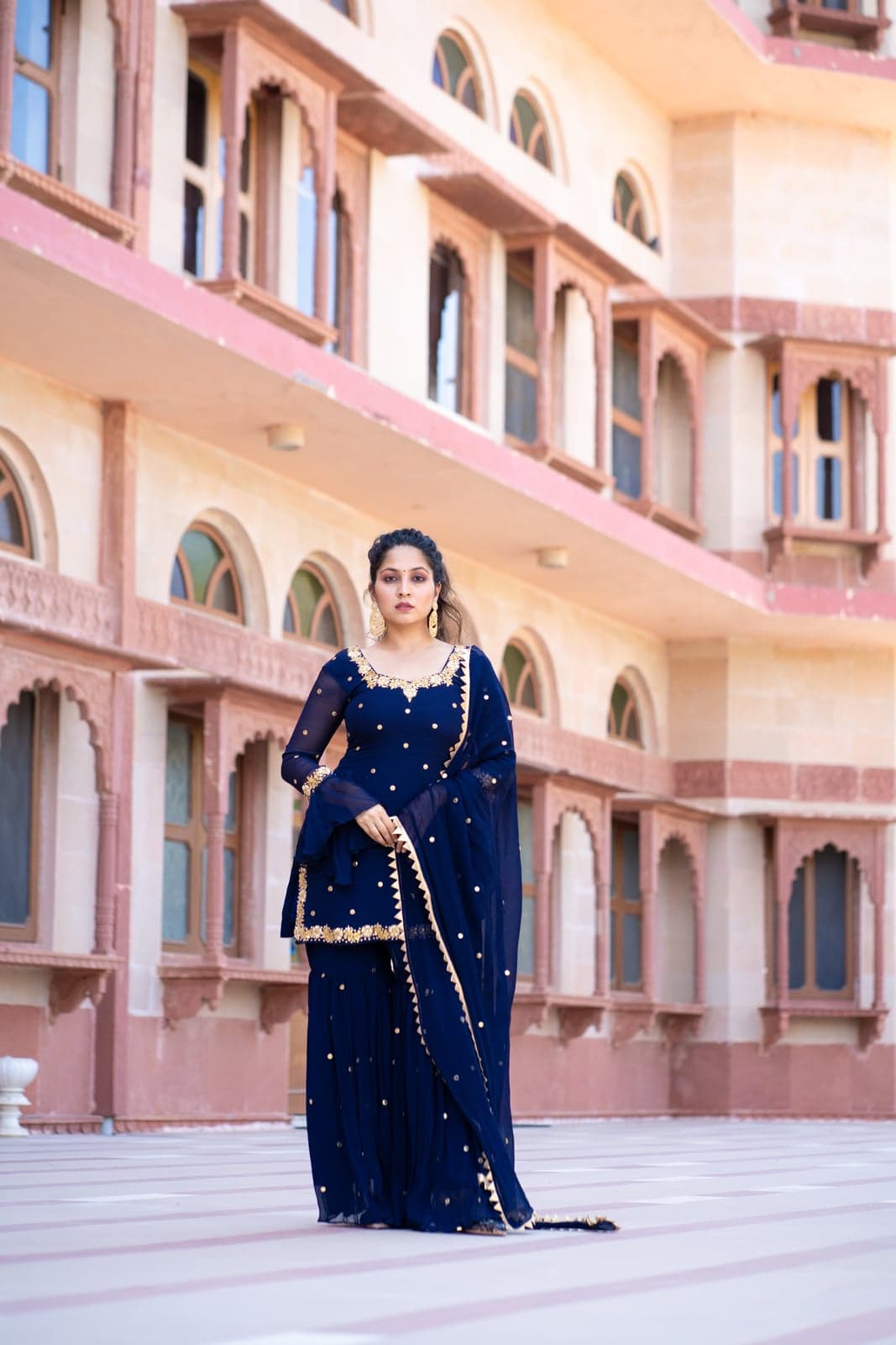 Navy chiffon kurta and gharara — gold mukaish dot embroidery with a zig-zag bordered dupatta, full-length view against a Rajasthani haveli facade.