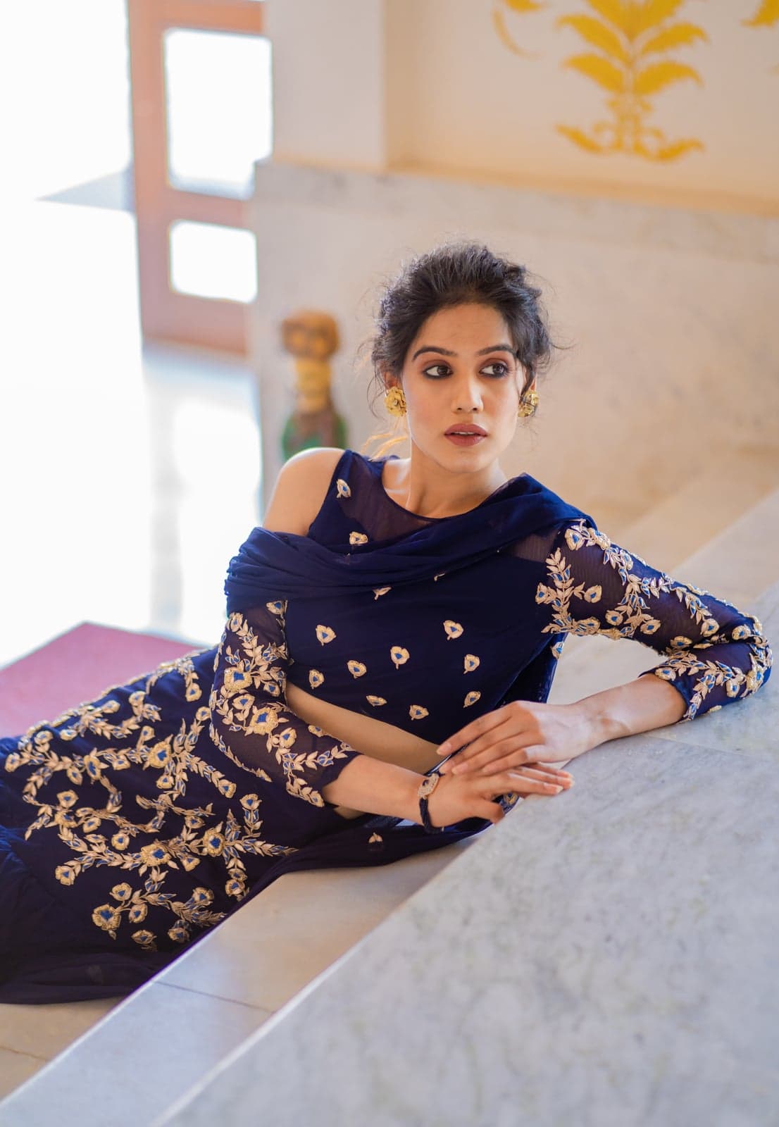 Navy lehenga reclining detail — gold-on-blue zardozi embroidery, sheer veil over the bodice, lit by lanterns in a marble hall.