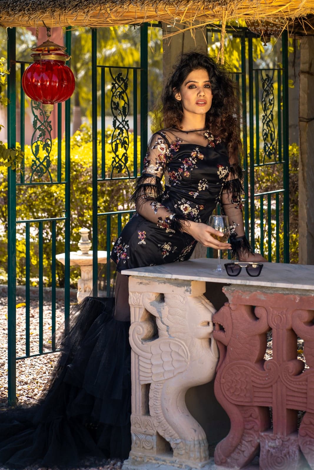 Black floral gown — close café-table portrait with a red glass lantern, tulle train trailing onto gravel.