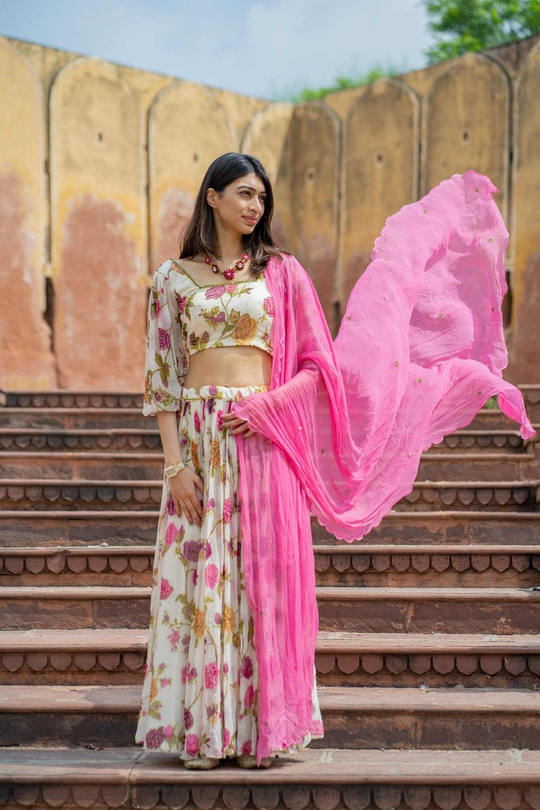 Cream silk crop blouse with a pink berry-and-leaf print and bell-balloon sleeves — paired with a hot-pink draped skirt, side portrait against a weathered Jaipur fort wall.