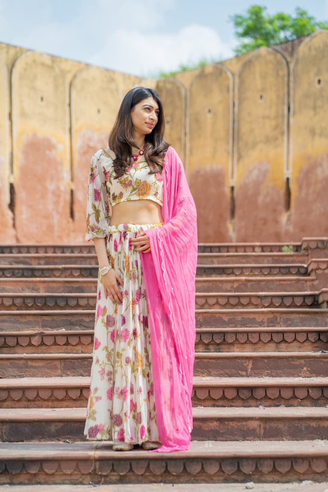 Cream berry-print crop with hot-pink draped skirt — facing-camera portrait against a faded ochre Jaipur wall.