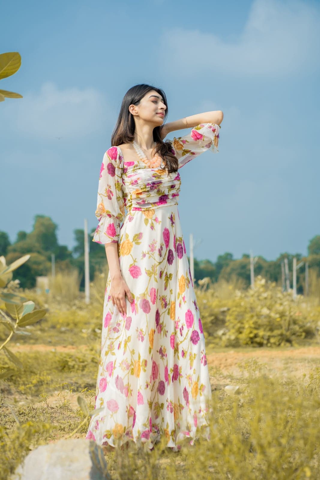 Ivory and pink rose-print chiffon maxi with cinched waist — full-length outdoor portrait in a sunlit field.