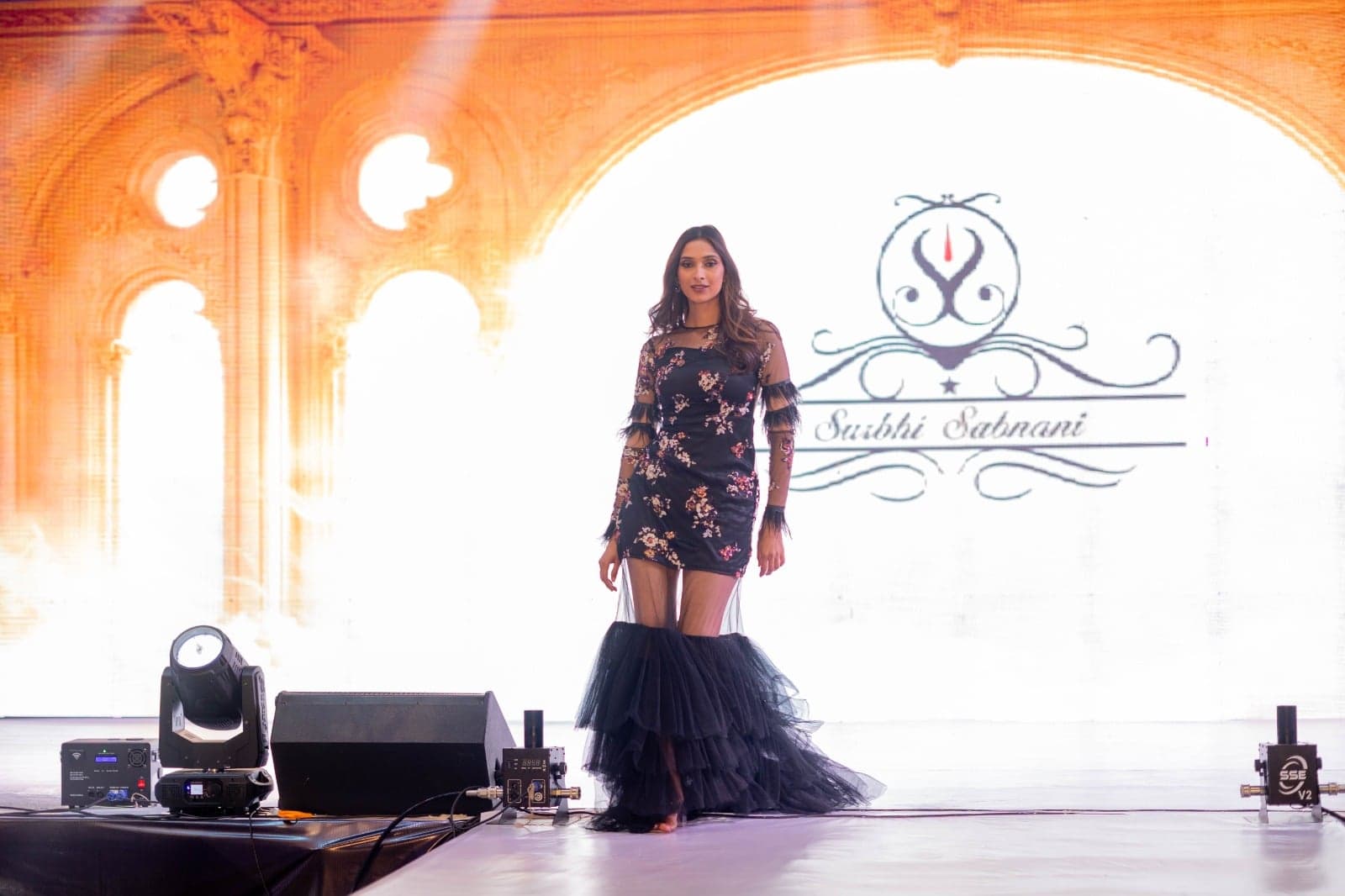 Black floral-embroidered mini dress with sheer mesh sleeves and a tiered tulle ruffle train — runway portrait beneath arched stage lights.