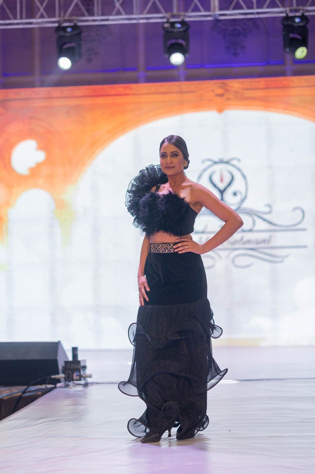 Black strapless dress with a feathered bodice, embellished belt, and tiered black-tulle ruffle skirt — runway portrait beneath stage lights.