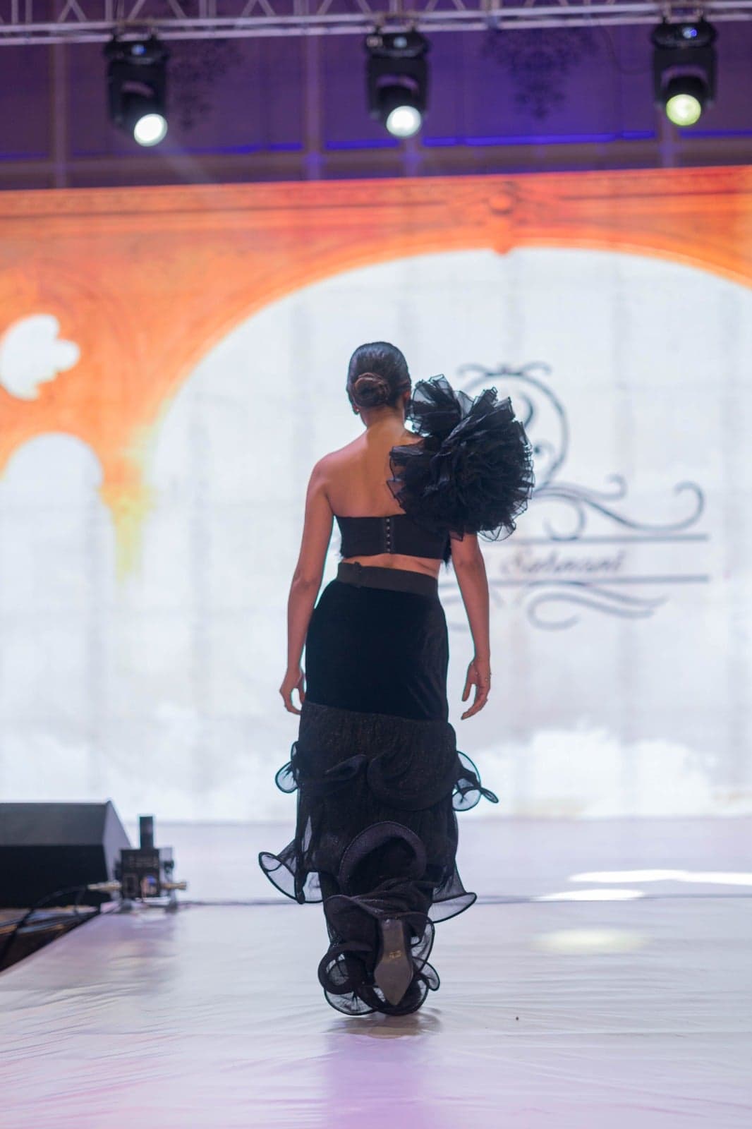 Black feathered-bodice gown — back view of the dramatic black-tulle pouf shoulder and tiered ruffle hem, mid-runway.