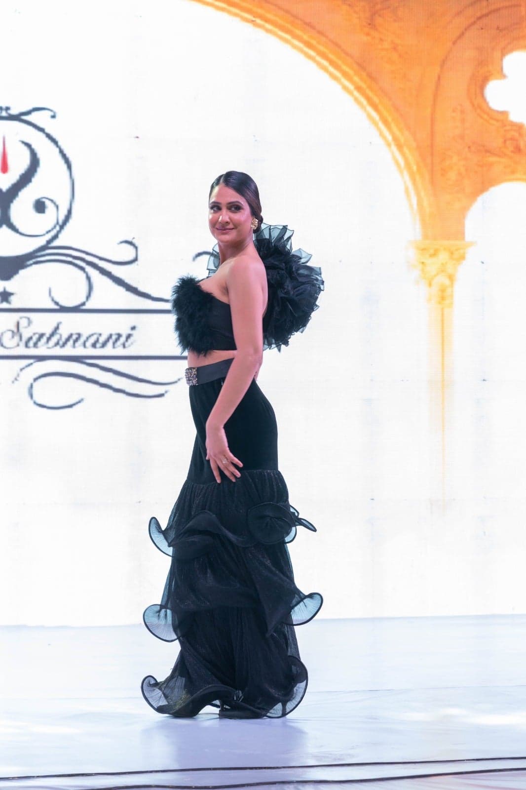 Black satin strapless gown with a feathered bodice and a high thigh-slit — runway portrait beneath arched stage lights.