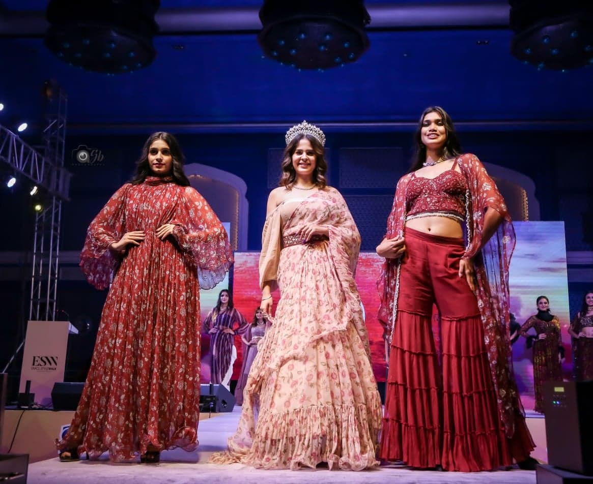 Trio runway portrait — coral-rust printed maxi, blush peach lehenga with sheer cape, and crimson tiered draped skirt with sheer printed cape.