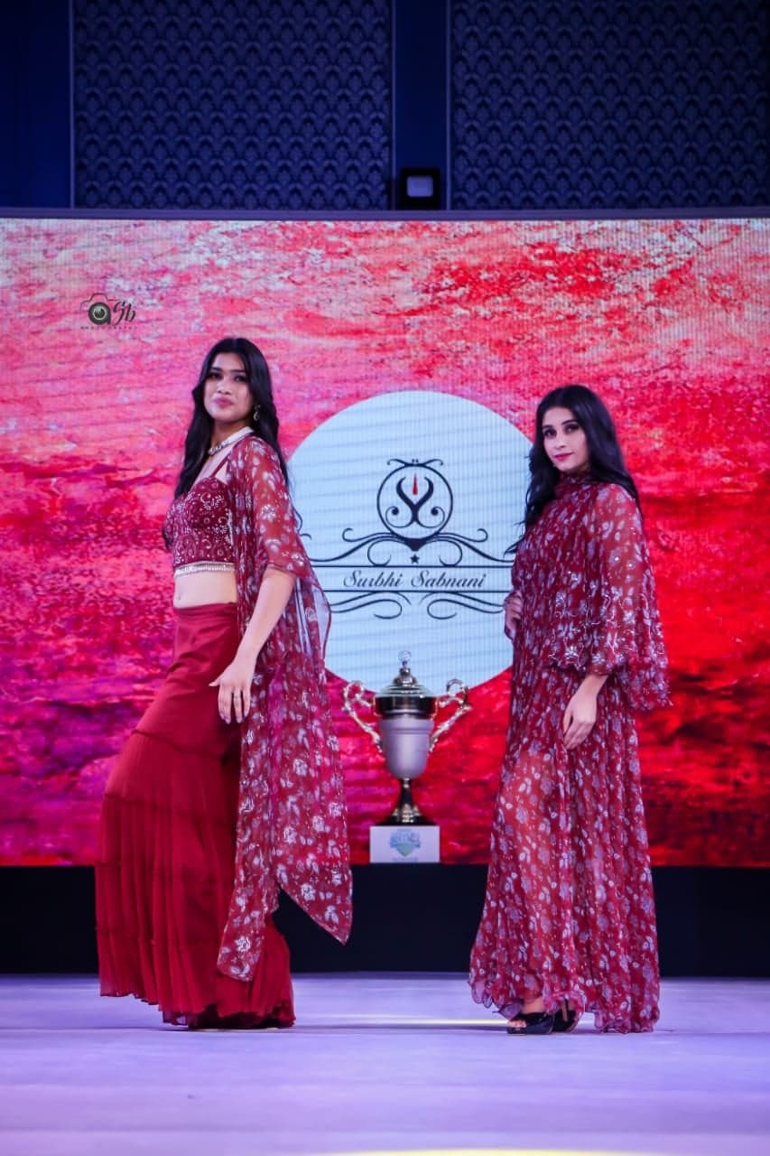 Two-model runway portrait — embellished crimson crop with printed sheer cape and crimson tiered skirt beside a dusty rose printed maxi-cape gown.