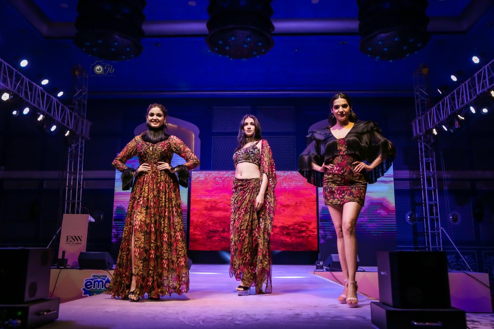 Trio runway portrait — burgundy-leaf printed maxi, sheer-cape crop-and-skirt set, and burgundy short dress with leaf-print ruffle wrap.