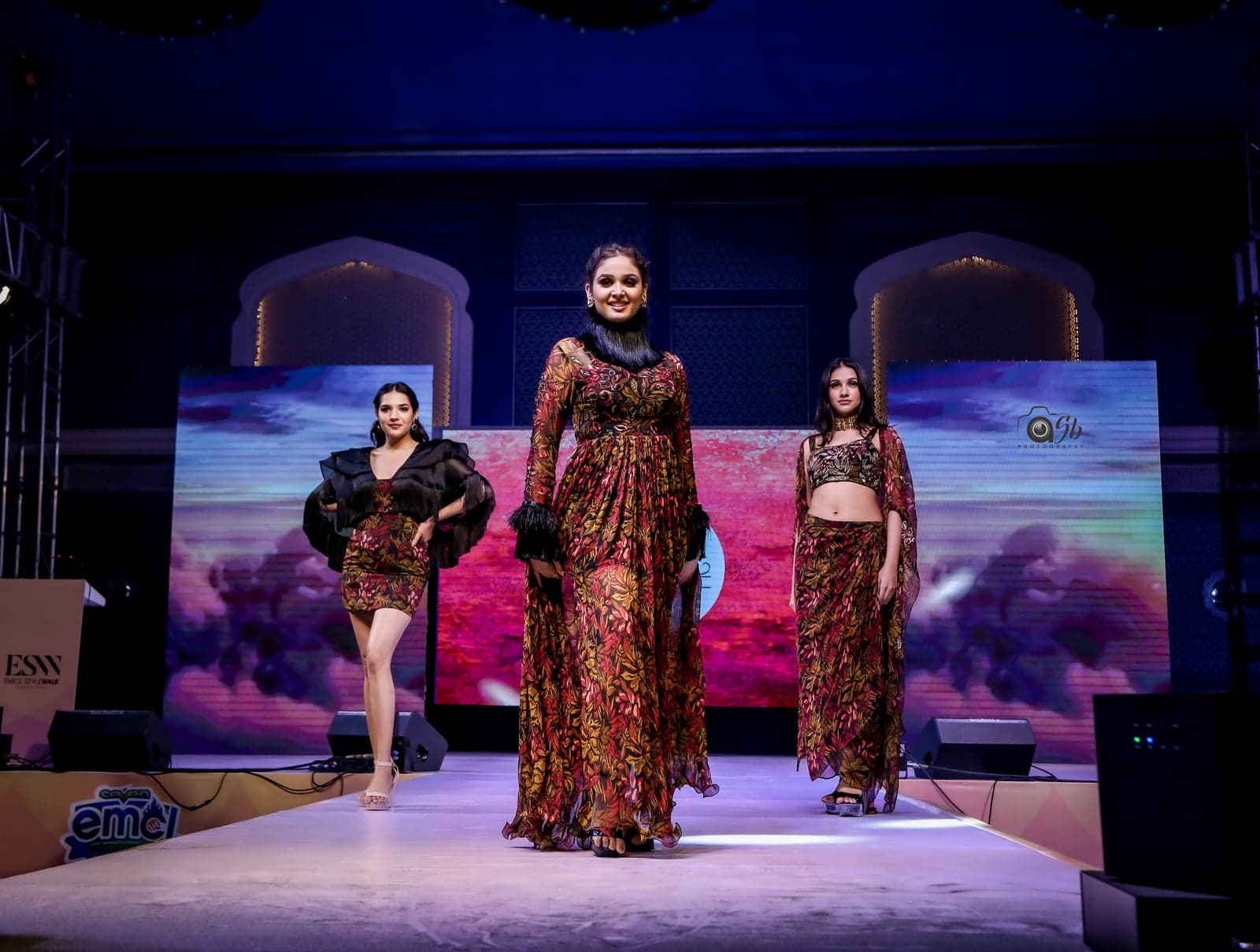Burgundy and gold leaf-print full-length gown with cinched waist and balloon sleeves — center of a trio runway portrait beneath stage lights.