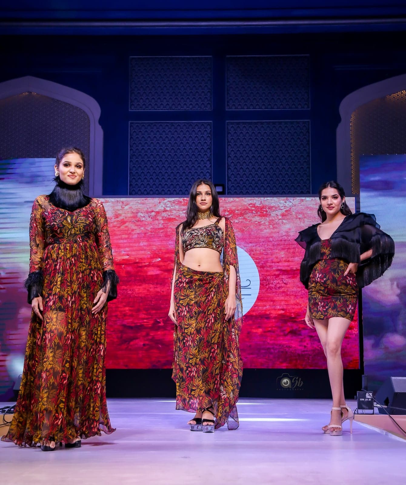 Trio runway portrait — burgundy leaf-print maxi, leaf-print crop-and-skirt, and leaf-print short dress with ruffle-cape, rust-toned backdrop.