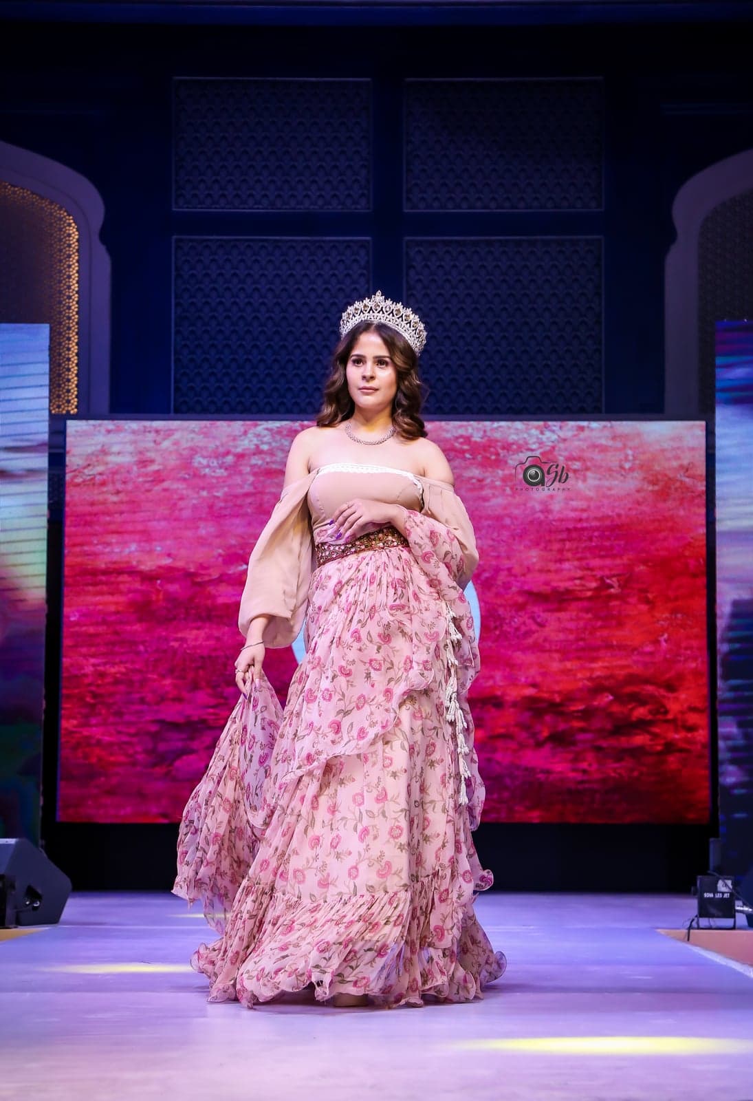 Blush peach maxi gown with a floral-rose print, off-shoulder bodice, balloon sleeves and a tiered ruffle skirt — runway portrait wearing a crown.