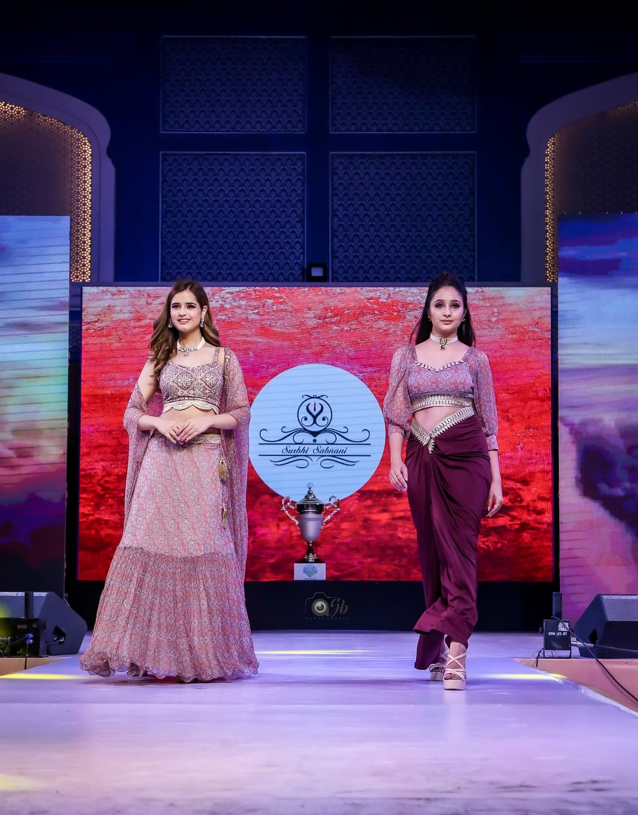 Two-model runway portrait — blush peach printed lehenga with sheer cape and maroon draped saree-skirt set, beneath stage chandelier.