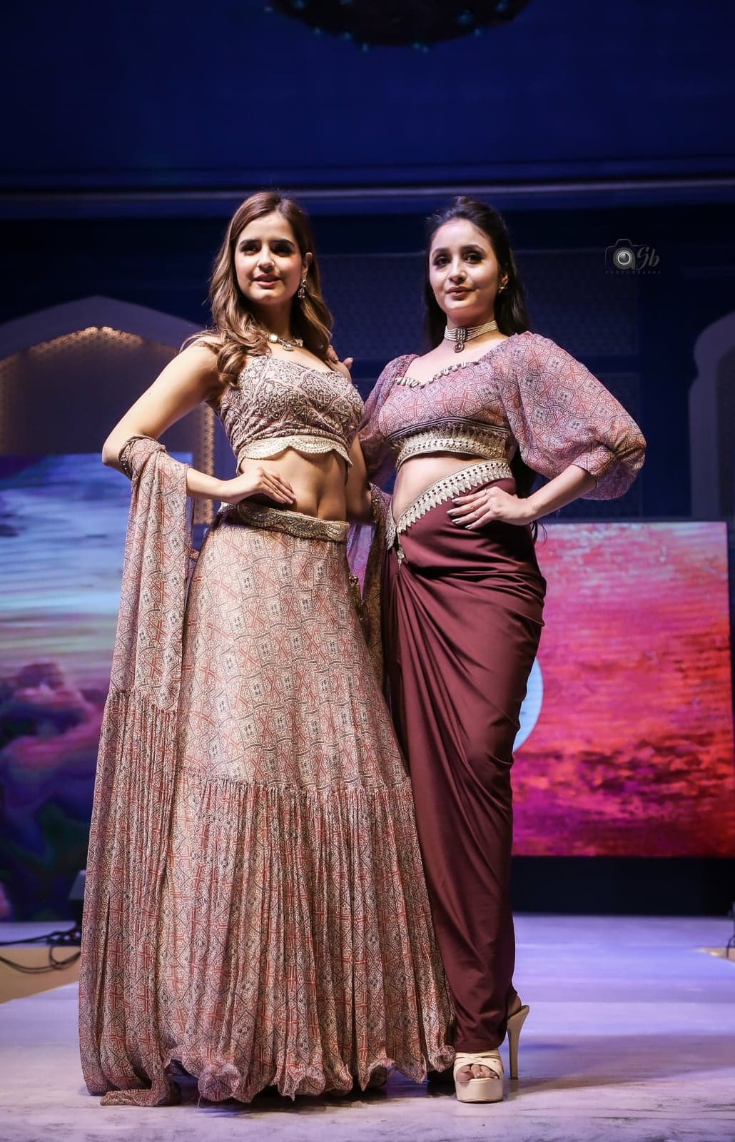 Two-model close runway portrait — blush peach lehenga and maroon dhoti skirt, embellished waist details in focus.