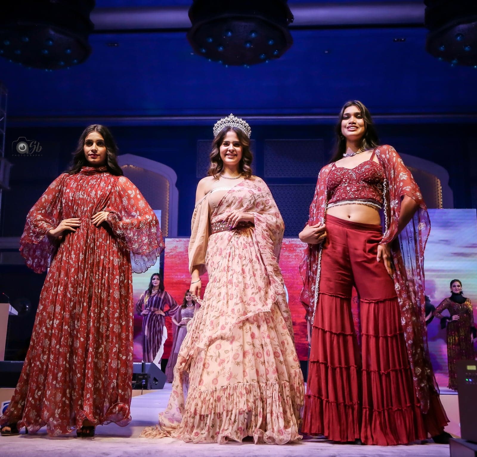 Trio runway portrait — burgundy leaf-print maxi at center, with blush peach lehenga and crimson tiered skirt with sheer cape to either side.