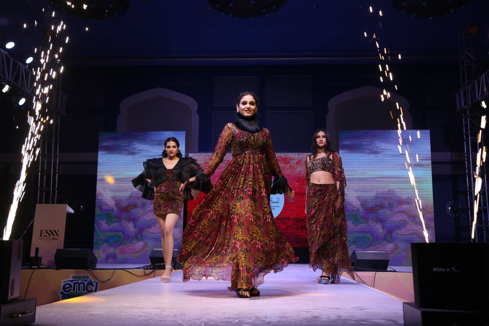 Trio runway portrait under pyro sparks — burgundy leaf-print collection in three silhouettes against the stage backdrop.