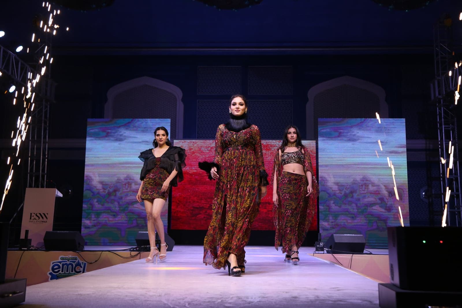 Trio runway portrait — burgundy leaf-print maxi, sheer-cape crop-and-skirt, and ruffle-shoulder short dress, beneath patterned wall backdrop.