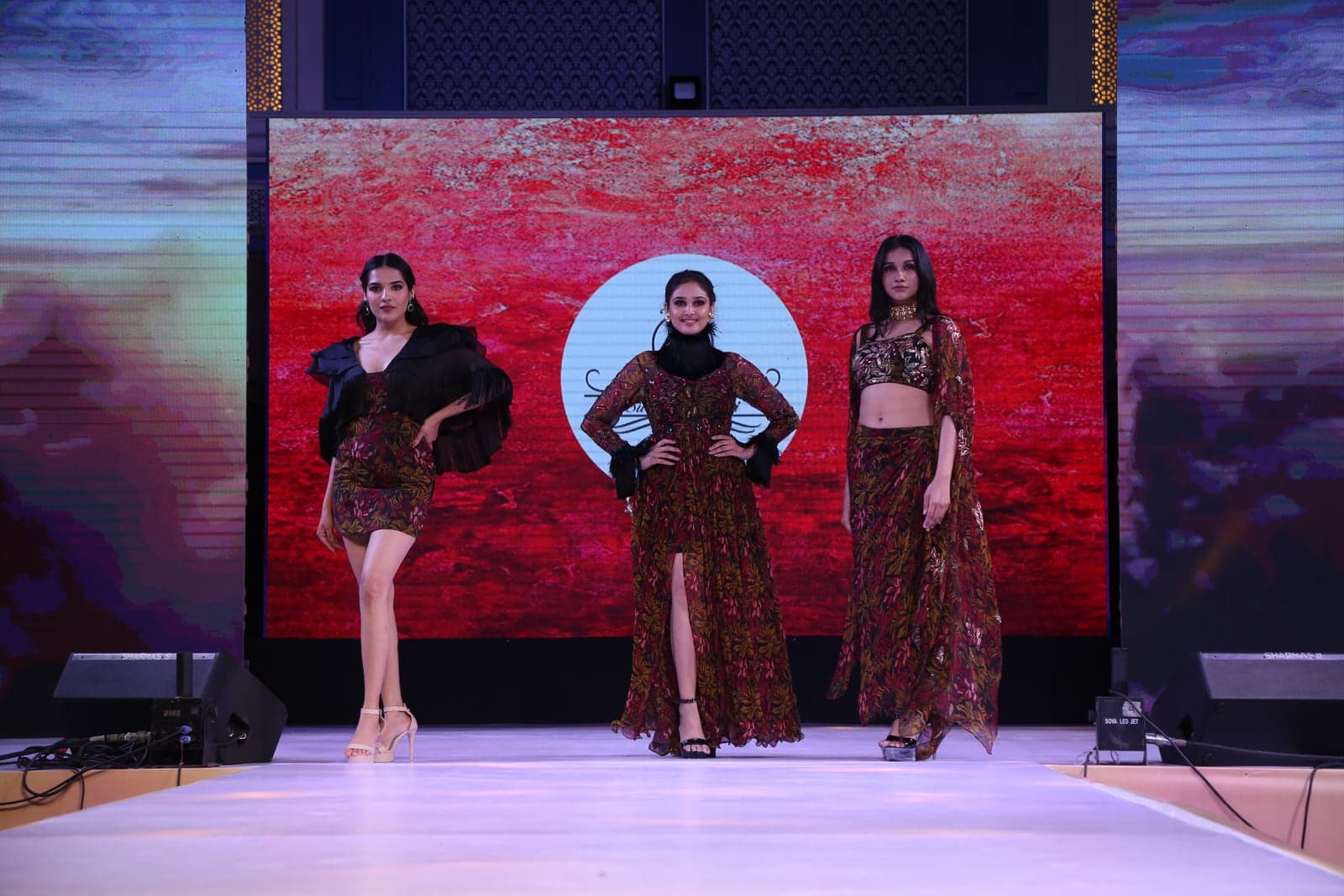 Trio runway portrait — leaf-print maxi, sheer-cape crop-and-skirt, and ruffle-shoulder short dress, all in the burgundy-leaf palette.