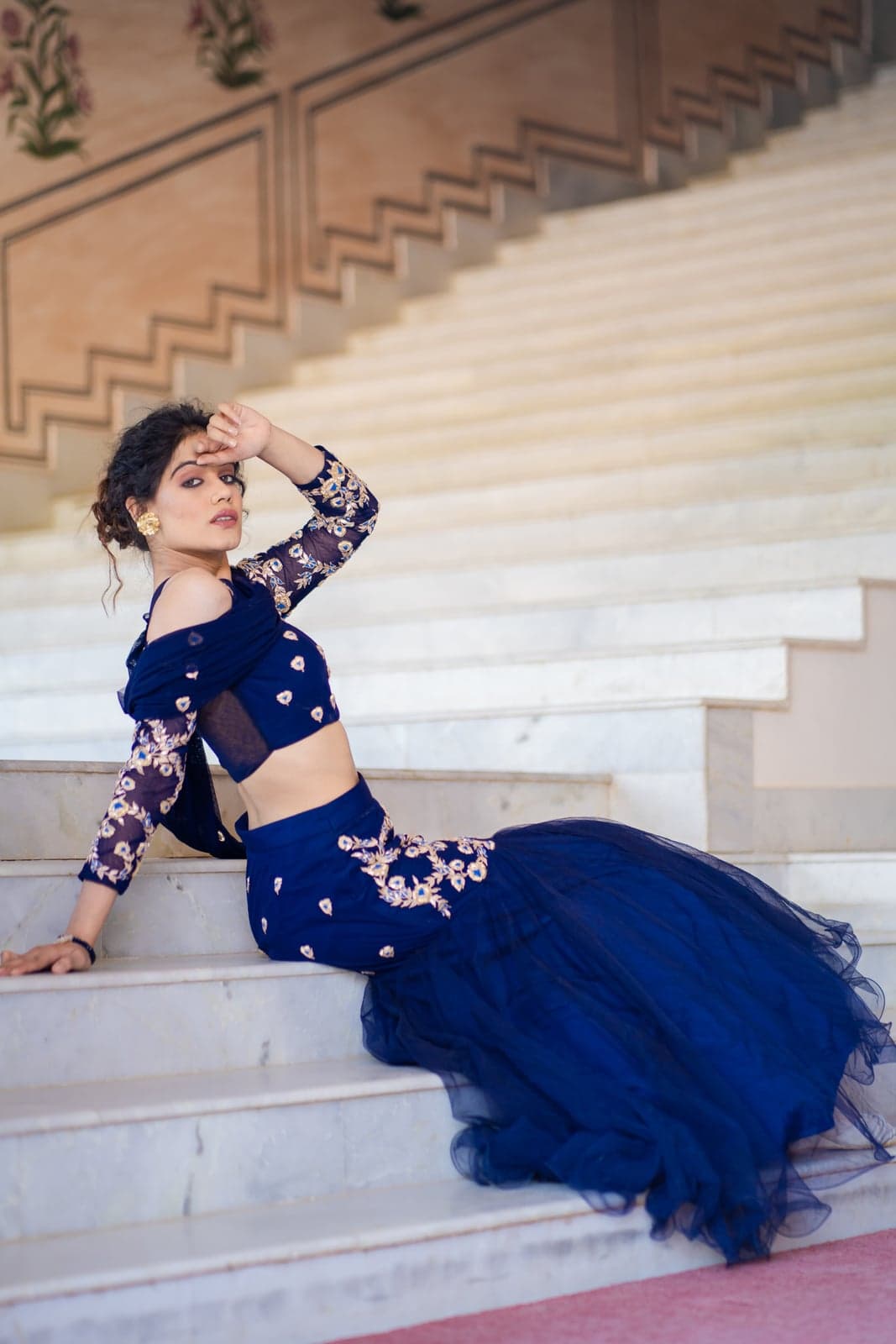 Sapphire blue lehenga with gold zardozi florals scattered across a sheer mesh blouse, paired with a flared tulle skirt — photographed on the marble steps of a Jaipur haveli.