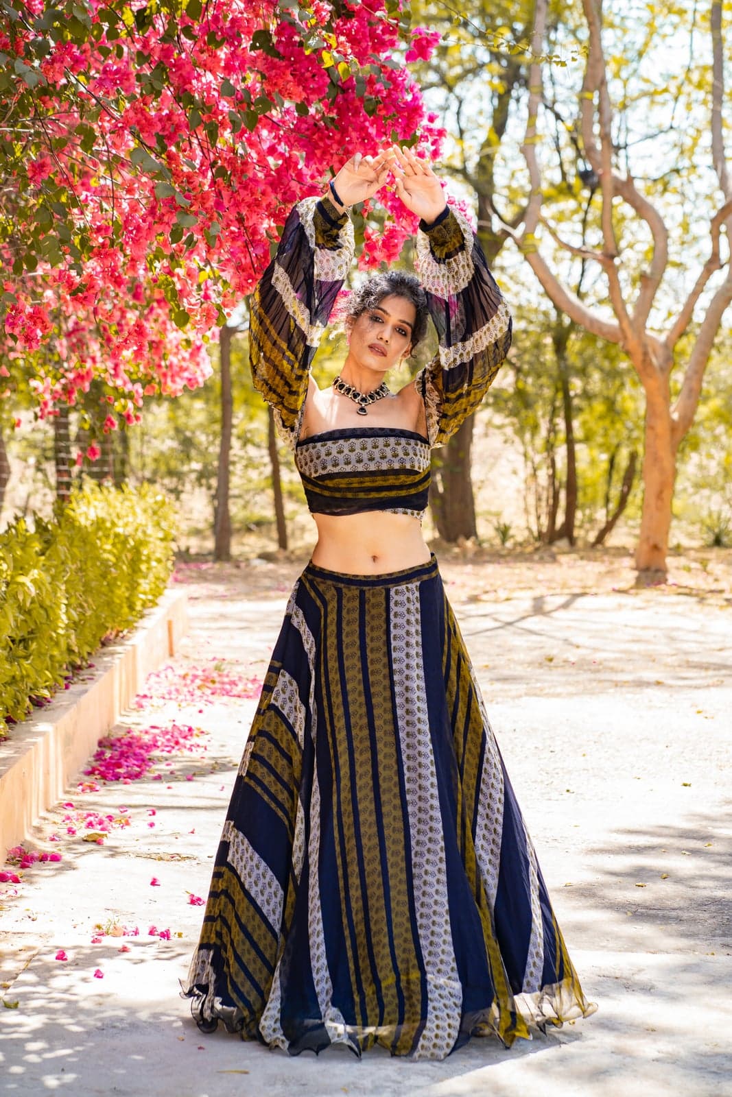 Indigo and ochre block-printed lehenga set with a striped pleated skirt and matching bell-sleeve blouse — photographed under a canopy of bougainvillea in bloom.