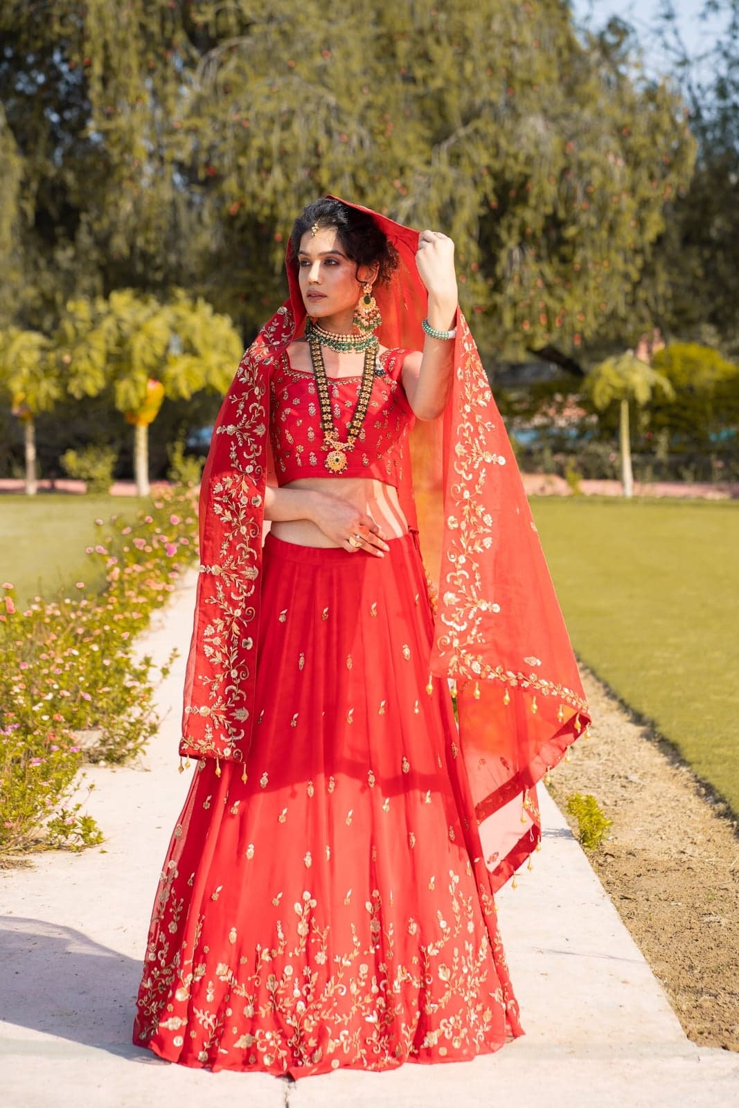 Scarlet red lehenga with gold-embroidered floral motifs and a sheer veil dupatta — Rajputana-bride styling, photographed in a garden at golden hour.