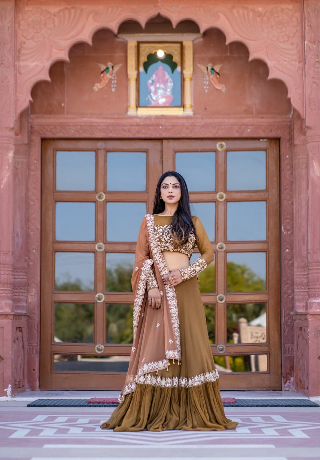 Caramel brown silk lehenga with a rich gold zardozi blouse and matching organza dupatta — framed within the cusped arch of a Rajasthani haveli doorway.