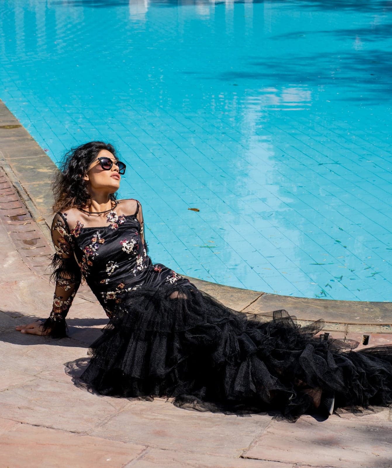 Black floral-embroidered gown with a tiered tulle train — sheer mesh bodice scattered with three-dimensional petalwork, photographed at the pool's edge.