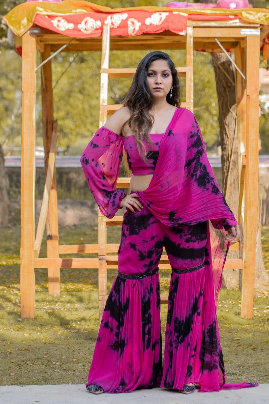 Magenta and black tie-dye chiffon gharara with an off-shoulder balloon-sleeve crop top and pleated wide-leg pants — photographed under a marigold-draped canopy.