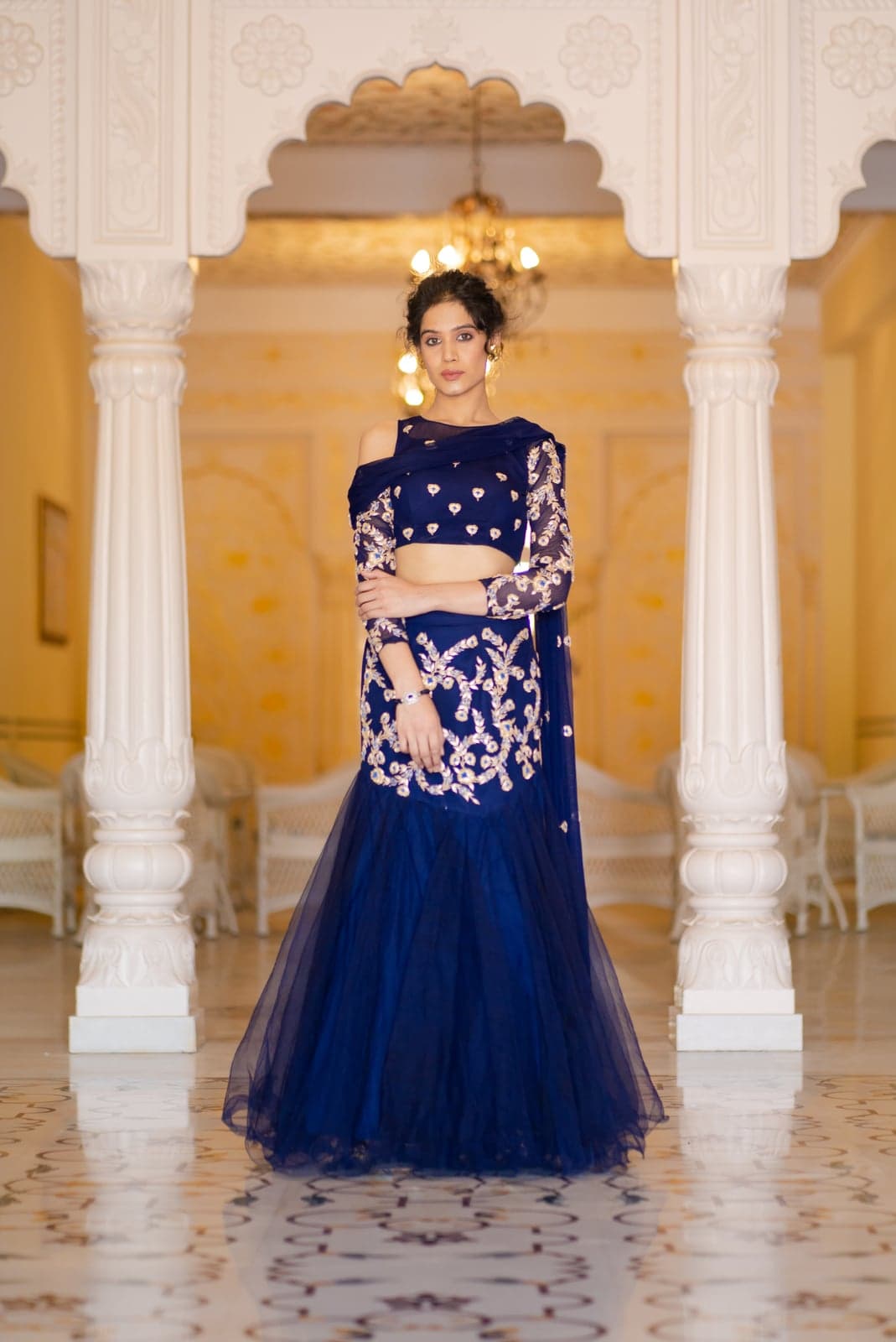 Sapphire blue lehenga in full silhouette — mermaid skirt, sheer floral-embroidered sleeves, photographed beneath the chandelier of a Jaipur palace hall.