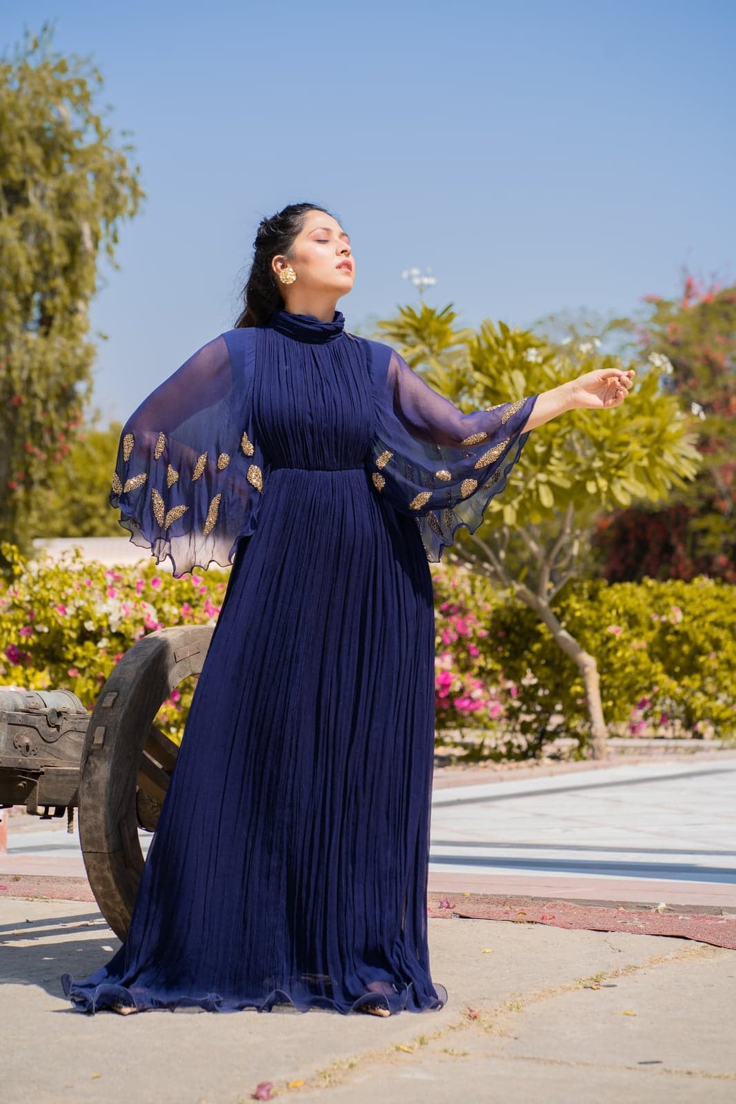 Navy pleated chiffon gown with a high collar and butterfly-cape sleeves embroidered in gold zardozi leaves — photographed in a sunlit Jaipur garden beside a vintage cannon.