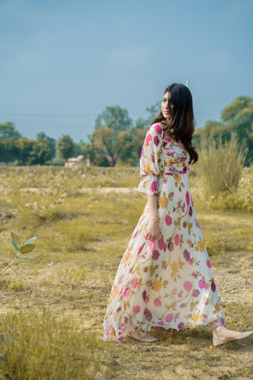 Ivory and pink rose-print chiffon maxi with a cinched waist and balloon sleeves — outdoor portrait in a sunlit clearing.