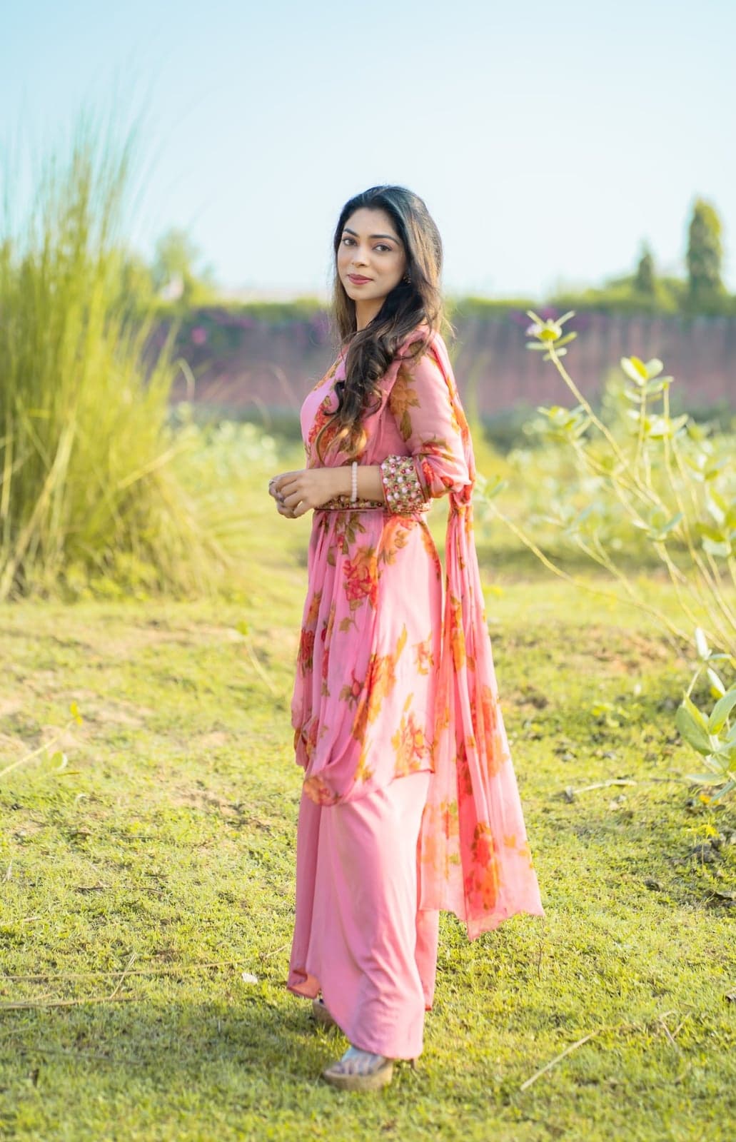 Pink chiffon kurta with orange rose-print balloon sleeves and matching dupatta — outdoor portrait beside tall grass tufts.