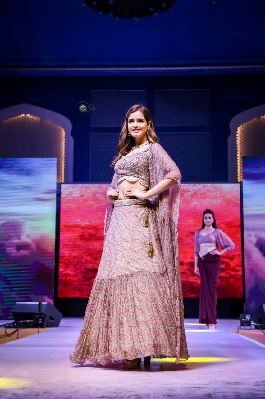 Blush peach printed lehenga with an embellished crop blouse and a sheer printed cape — runway portrait against the red-and-blue sunset backdrop.